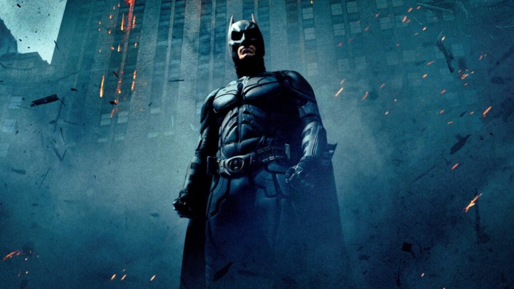 Batman em pé diante de prédios e destroços em cena da trilogia de Christopher Nolan