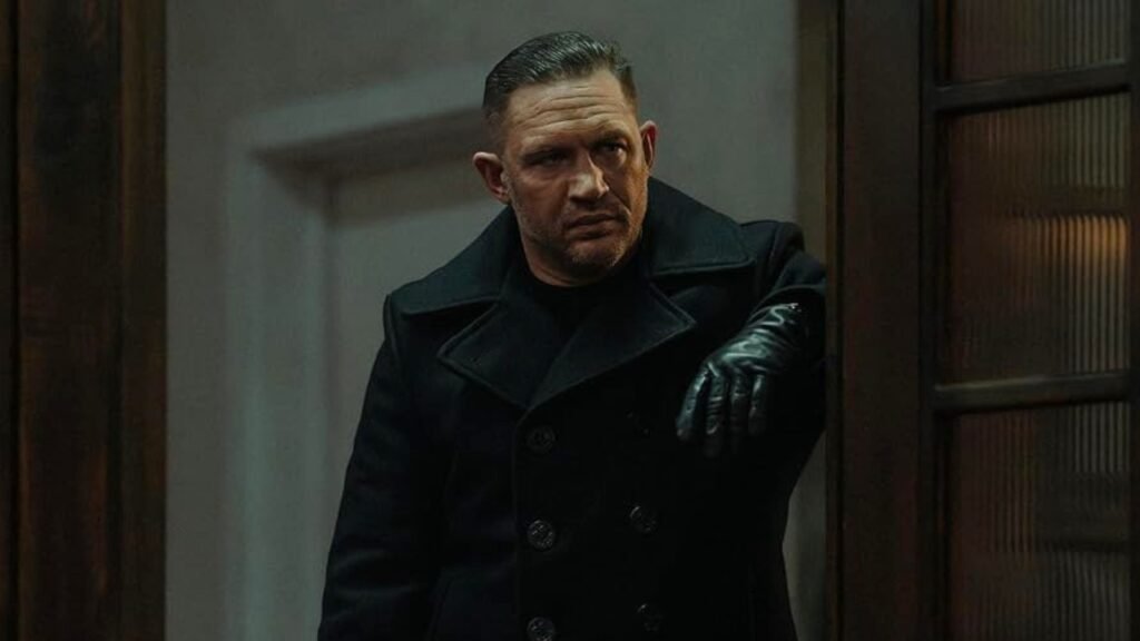 Harry Da Souza, personagem de Tom Hardy, apoia o braço enluvado na porta enquanto observa alguém em cena de Terra da Máfia.